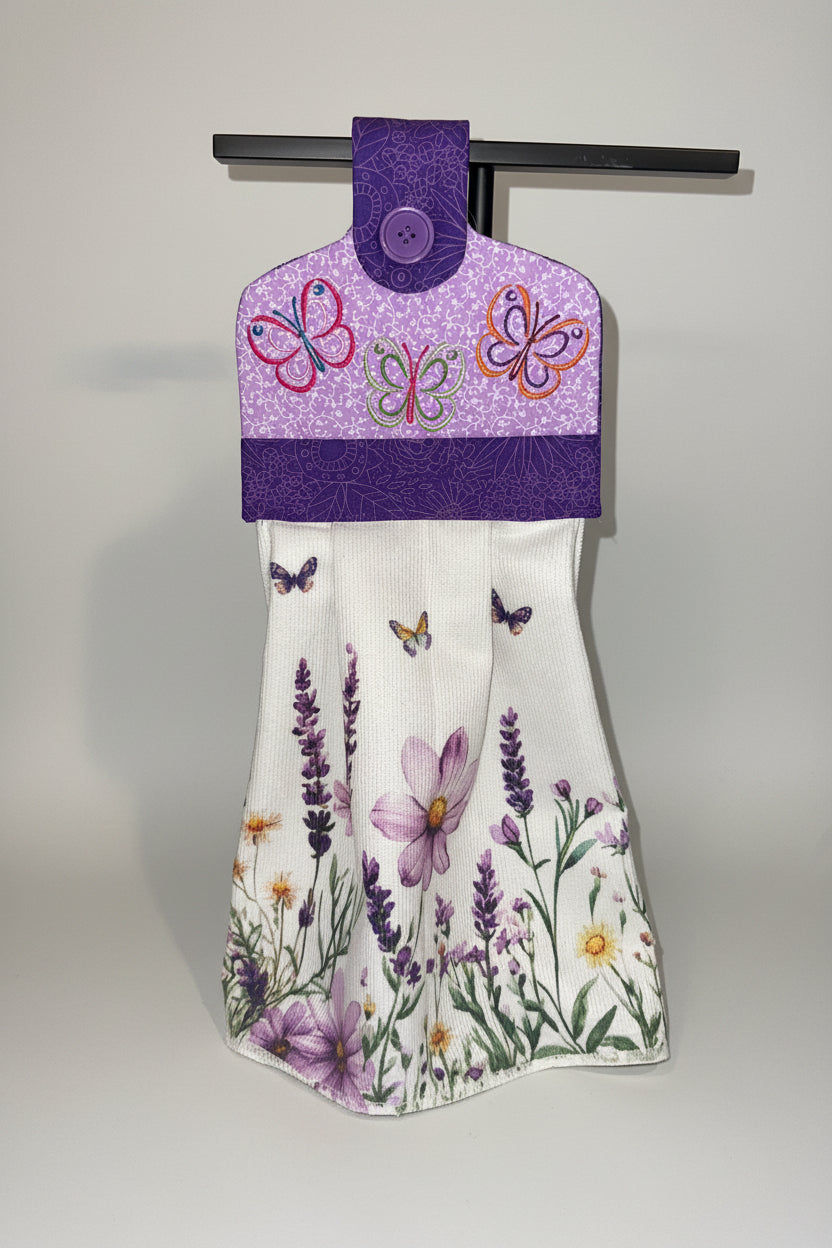 A hanging kitchen towel featuring a watercolor style print of purple flowers and butterflies. The top section is purple with a quilted texture, embroidered butterflies, a purple patterned fabric border, with a button closure.