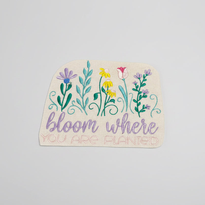 Embroidered design with flowers and text 'bloom where you are planted' on a beige background