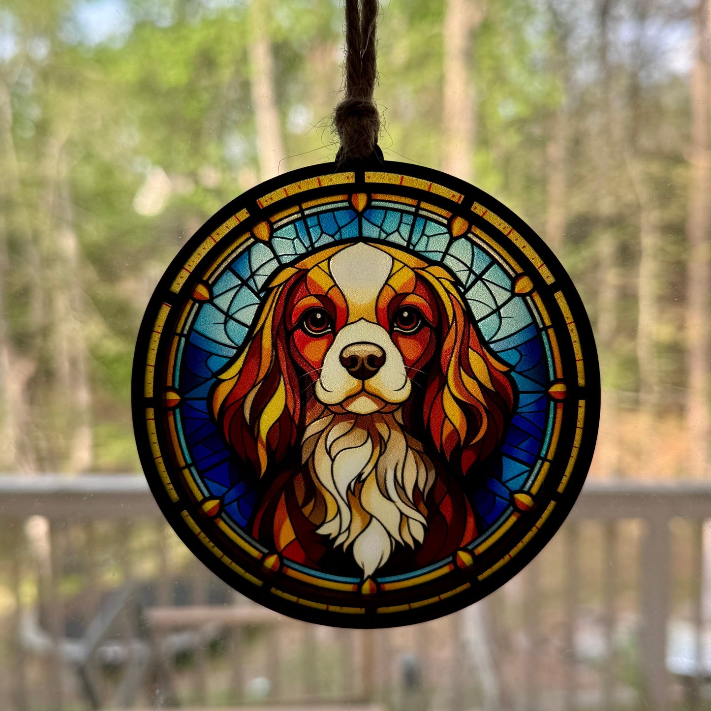 Stained glass looking sun catcher with a Cavalier King Charles dog design hanging in a window with a wooded background outdoors