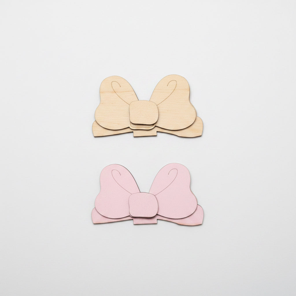 Two bow-shaped MDF cutouts, one unpainted and one pink, on a brown surface