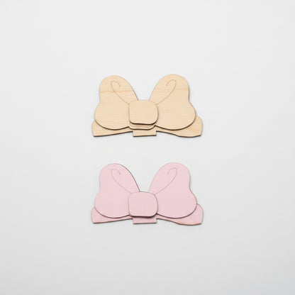Two bow-shaped MDF cutouts, one unpainted and one pink, on a brown surface