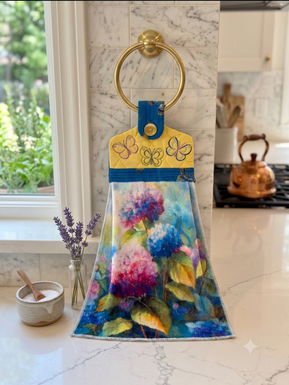 A hanging kitchen towel featuring a watercolor style print of blue and pink hydrangeas. The top section is yellow with a quilted texture, embroidered butterflies, a blue patterned fabric border, and a button closure.