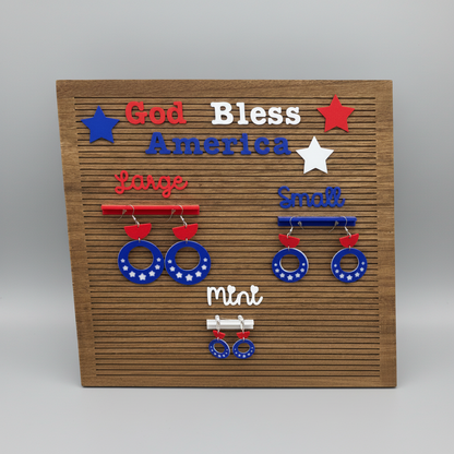 Patriotic Earrings of different sizes on a wood board that says God Bless America