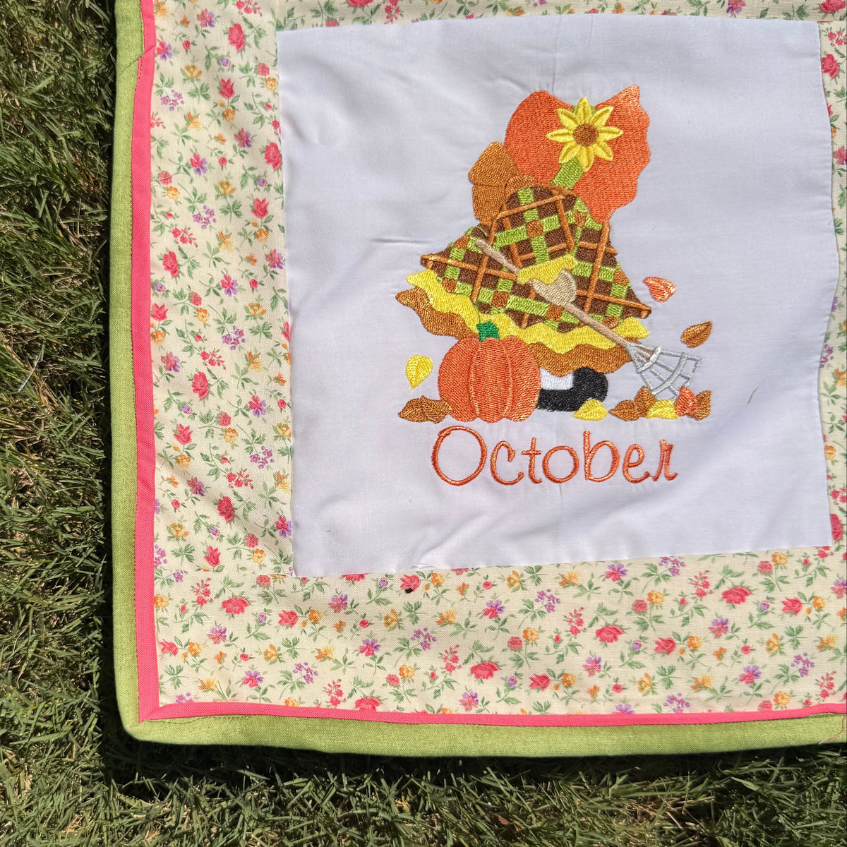 A close-up of a handmade quilted blanket or baby blanket laid on green grass outdoors. The blanket features set of Sunbonnet Sue Illustrations for each month of the year.