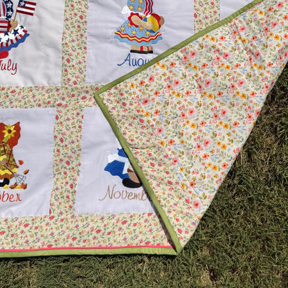 A close-up of a handmade quilted blanket or baby blanket laid on green grass outdoors. The blanket features set of Sunbonnet Sue Illustrations for each month of the year.