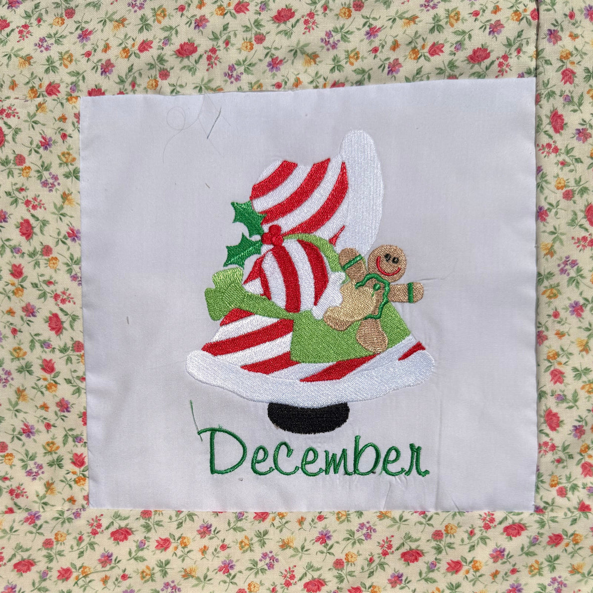 A close-up of a handmade quilted blanket or baby blanket laid on green grass outdoors. The blanket features set of Sunbonnet Sue Illustrations for each month of the year.