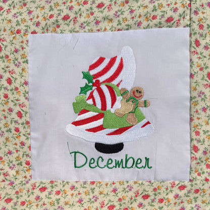 A close-up of a handmade quilted blanket or baby blanket laid on green grass outdoors. The blanket features set of Sunbonnet Sue Illustrations for each month of the year.