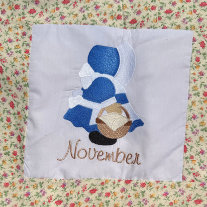 A close-up of a handmade quilted blanket or baby blanket laid on green grass outdoors. The blanket features set of Sunbonnet Sue Illustrations for each month of the year.