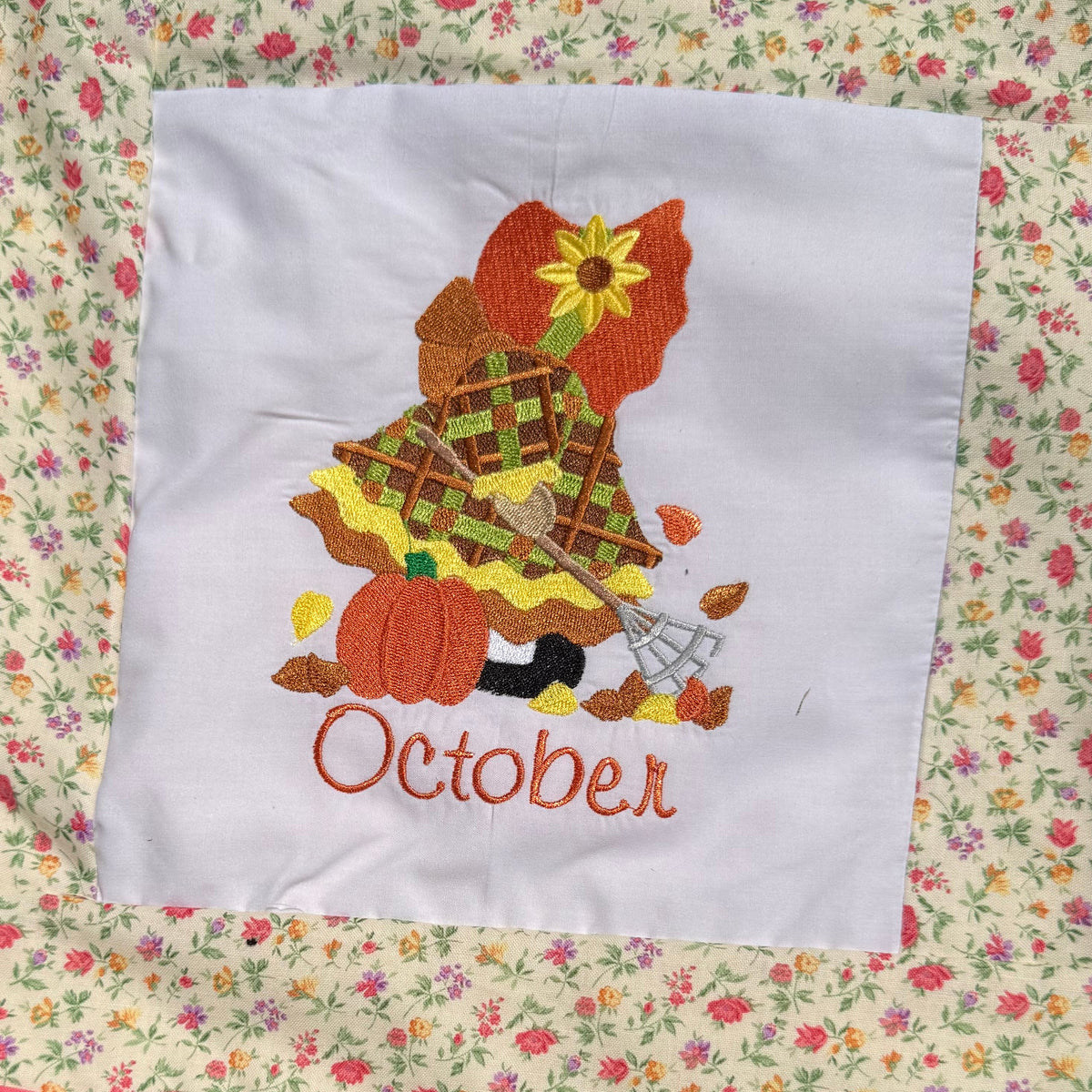 A close-up of a handmade quilted blanket or baby blanket laid on green grass outdoors. The blanket features set of Sunbonnet Sue Illustrations for each month of the year.