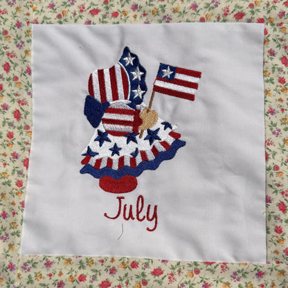 A close-up of a handmade quilted blanket or baby blanket laid on green grass outdoors. The blanket features set of Sunbonnet Sue Illustrations for each month of the year.