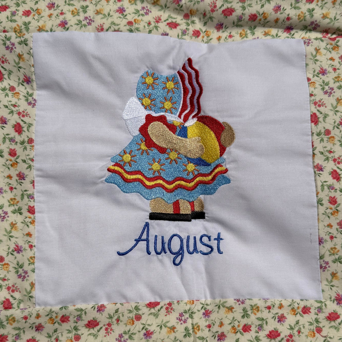 A close-up of a handmade quilted blanket or baby blanket laid on green grass outdoors. The blanket features set of Sunbonnet Sue Illustrations for each month of the year.