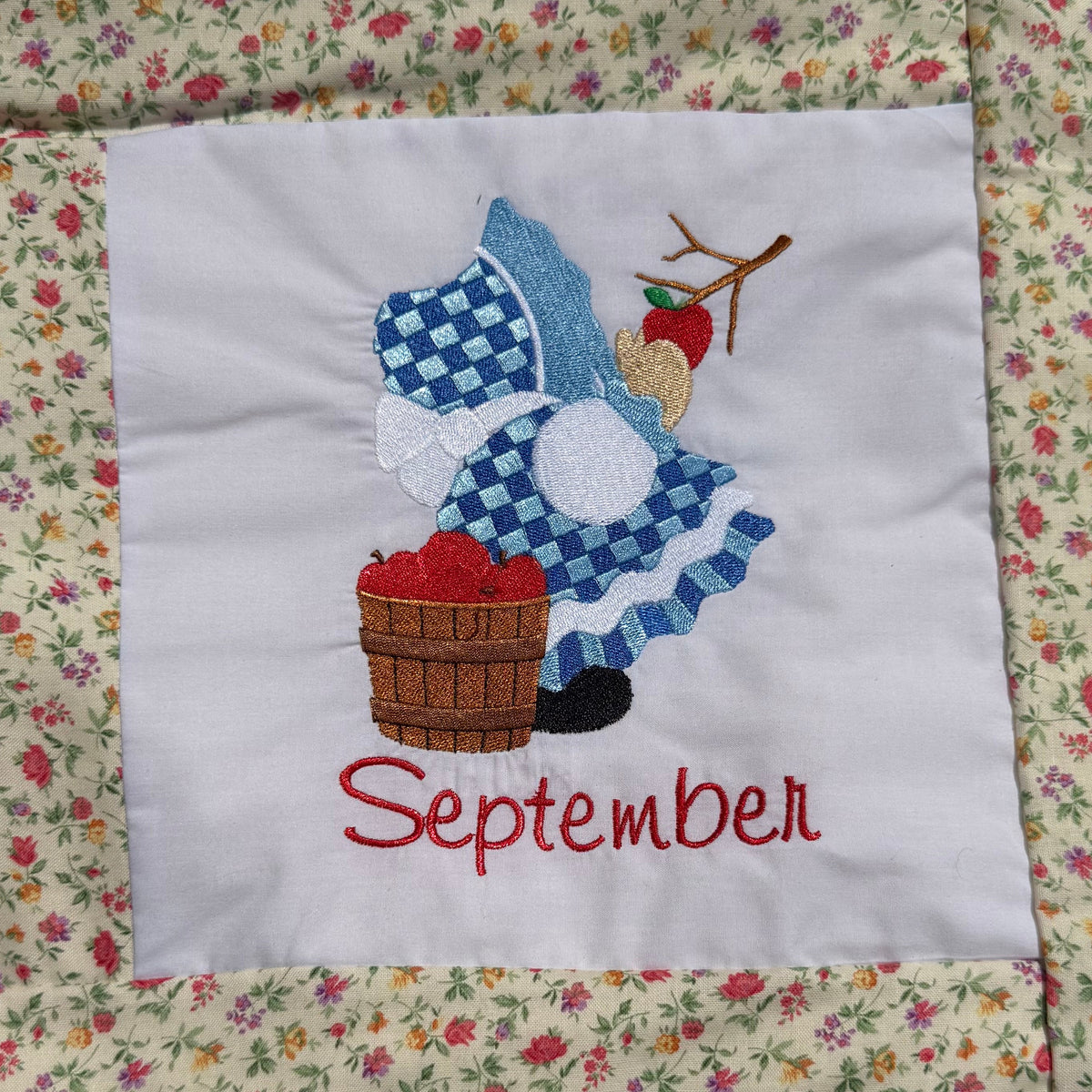 A close-up of a handmade quilted blanket or baby blanket laid on green grass outdoors. The blanket features set of Sunbonnet Sue Illustrations for each month of the year.