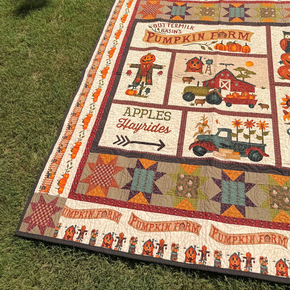 A large patchwork quilt laid flat on green grass outdoors, featuring autumn-themed designs including pumpkins, a barn, a truck, and text such as 'Pumpkin Farm' and 'Apples Hayrides.' Two people are standing in the background on the lawn.