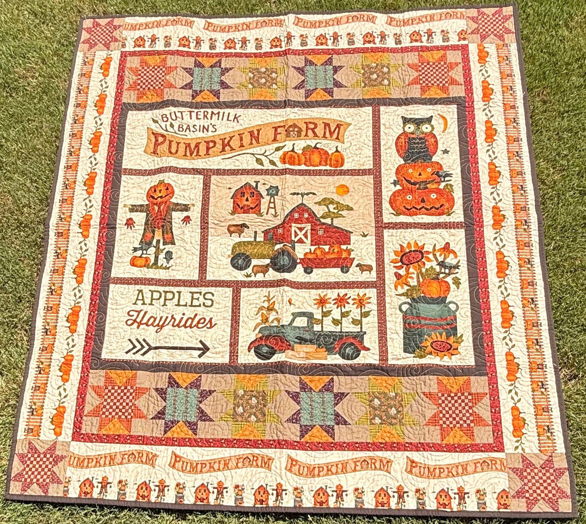 A square patchwork quilt laid flat on a green grass lawn. The quilt features a rustic, multi-colored patchwork design with various folk-art style fall pumpkin farm illustrations including a scarecrow, an old truck, a pumpkin stack and more.