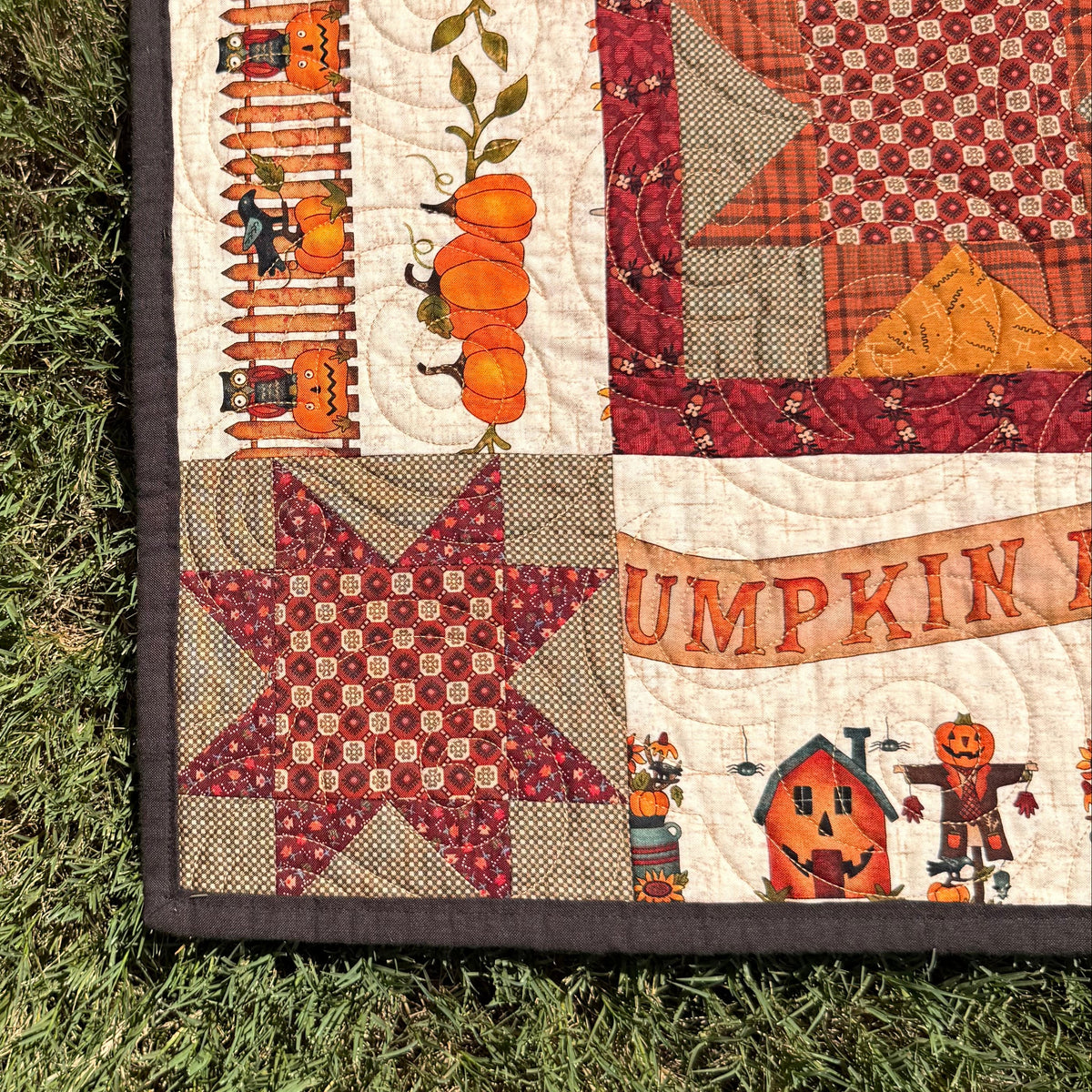 An autumn-themed quilted blanket or quilt laid out on green grass, featuring orange pumpkins, a red star patch, and a pumpkin patch illustration.