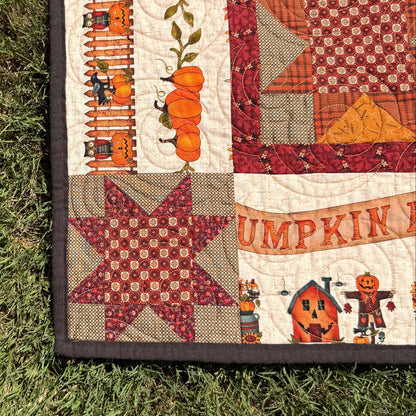 An autumn-themed quilted blanket or quilt laid out on green grass, featuring orange pumpkins, a red star patch, and a pumpkin patch illustration.