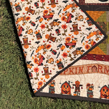 A folded quilt with a colorful autumn-themed print and a patterned backing, laid on green grass outdoors.