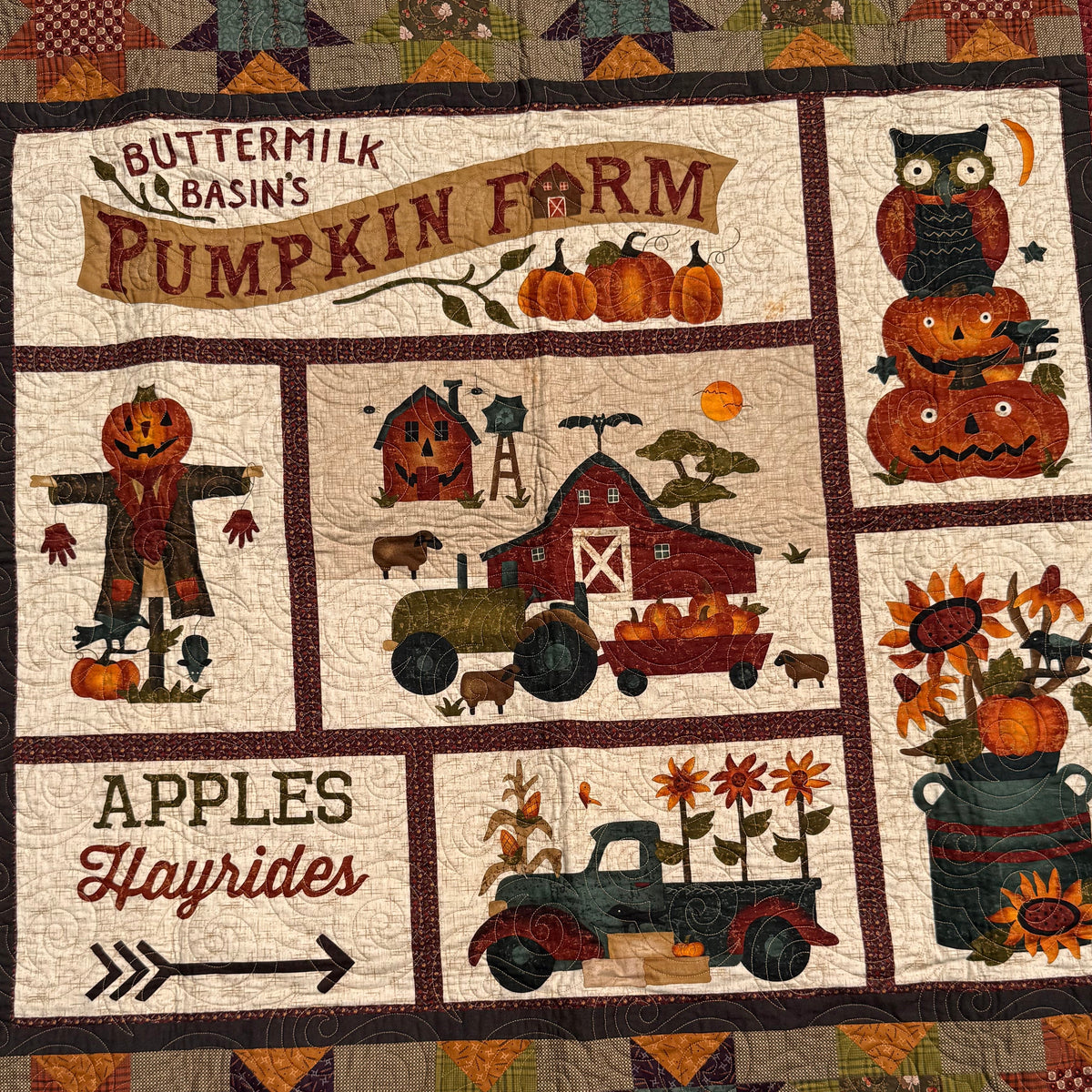 A handmade autumn-themed quilt laid flat outdoors on grass. The quilt features a cream background with multiple panels depicting fall harvest motifs including pumpkins, scarecrows, a barn, a tractor, and text such as 'Apples Hayrides' and 'Buttermilk Basin's Pumpkin Farm'.