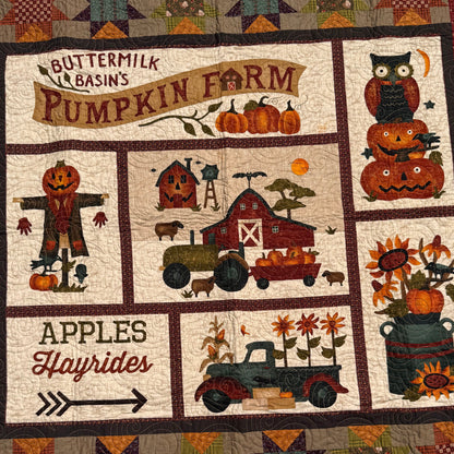 A handmade autumn-themed quilt laid flat outdoors on grass. The quilt features a cream background with multiple panels depicting fall harvest motifs including pumpkins, scarecrows, a barn, a tractor, and text such as 'Apples Hayrides' and 'Buttermilk Basin's Pumpkin Farm'.