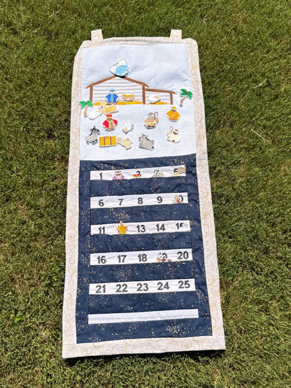 A handmade fabric advent calendar on a green grass lawn. The calendar has a nativity scene on the top section with nativity themed characters and decorations the attach with velcro and fit in the numbered pockets in the lower section.
