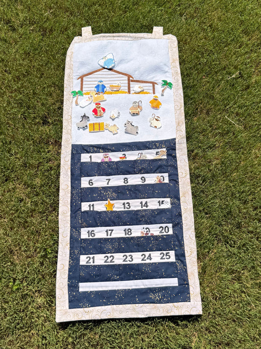 A handmade fabric advent calendar on a green grass lawn. The calendar has a nativity scene on the top section with nativity themed characters and decorations the attach with velcro and fit in the numbered pockets in the lower section.