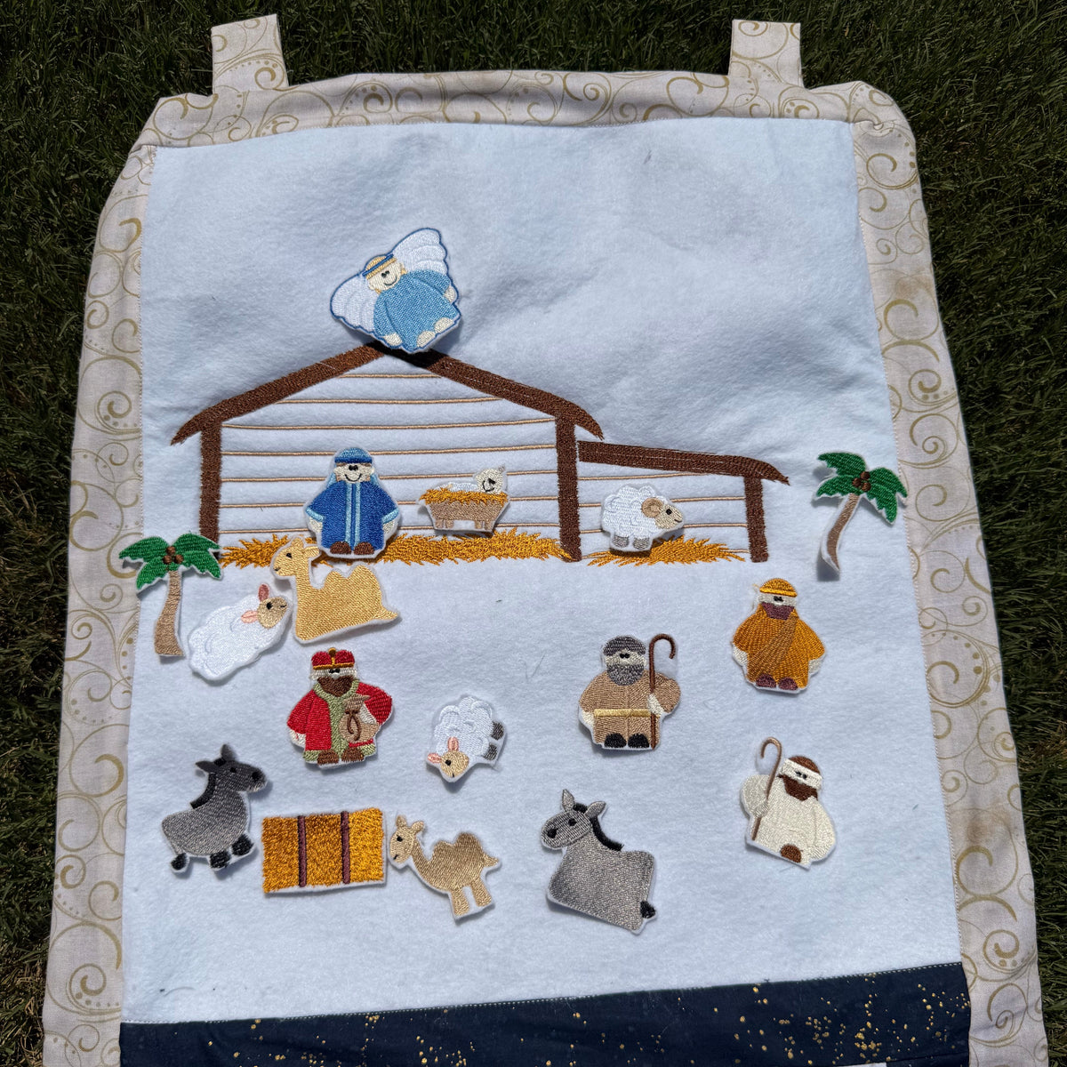 A handmade fabric advent calendar on a green grass lawn. The calendar has a nativity scene on the top section with nativity themed characters and decorations the attach with velcro and fit in the numbered pockets in the lower section.