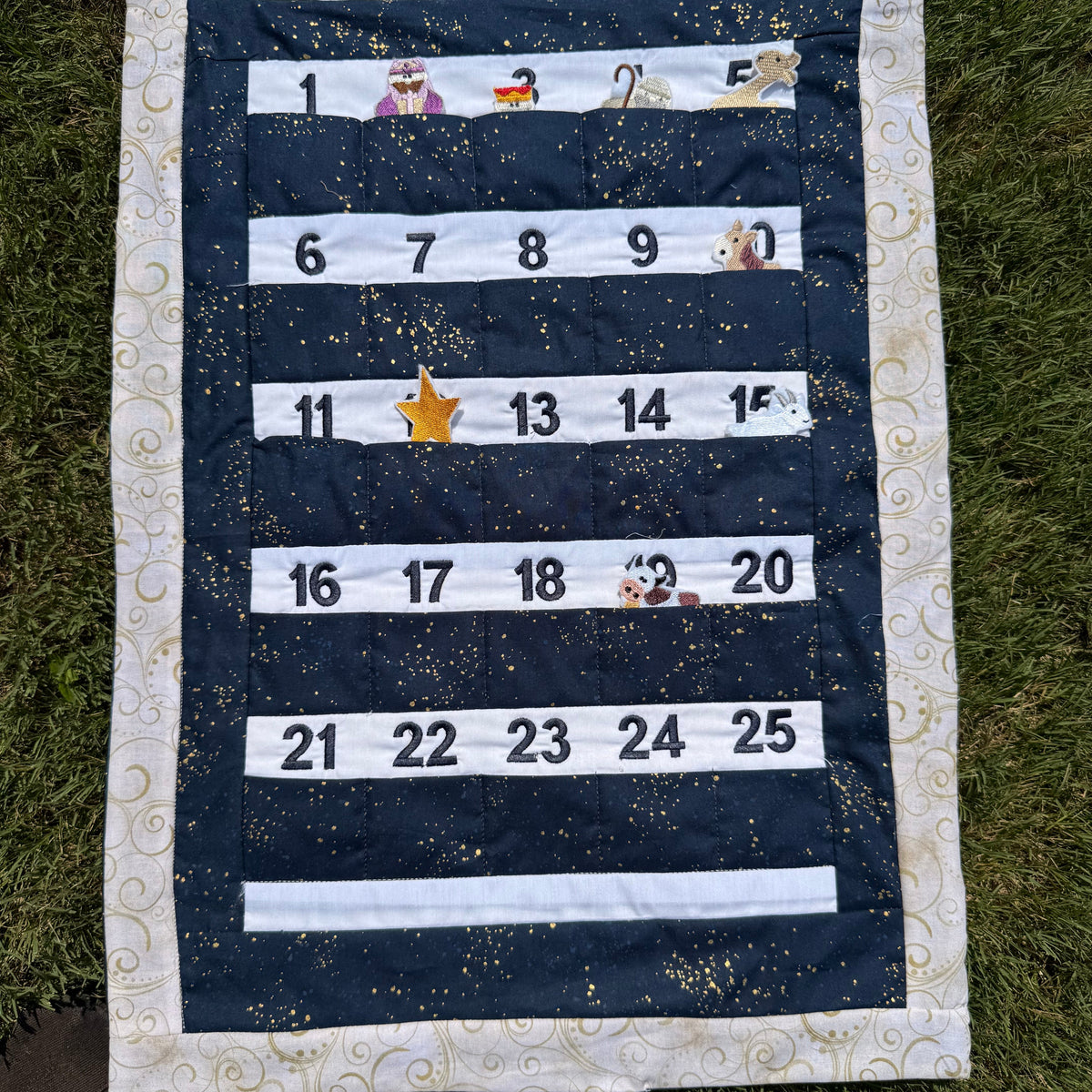 A handmade fabric advent calendar on a green grass lawn. The calendar has a nativity scene on the top section with nativity themed characters and decorations the attach with velcro and fit in the numbered pockets in the lower section.