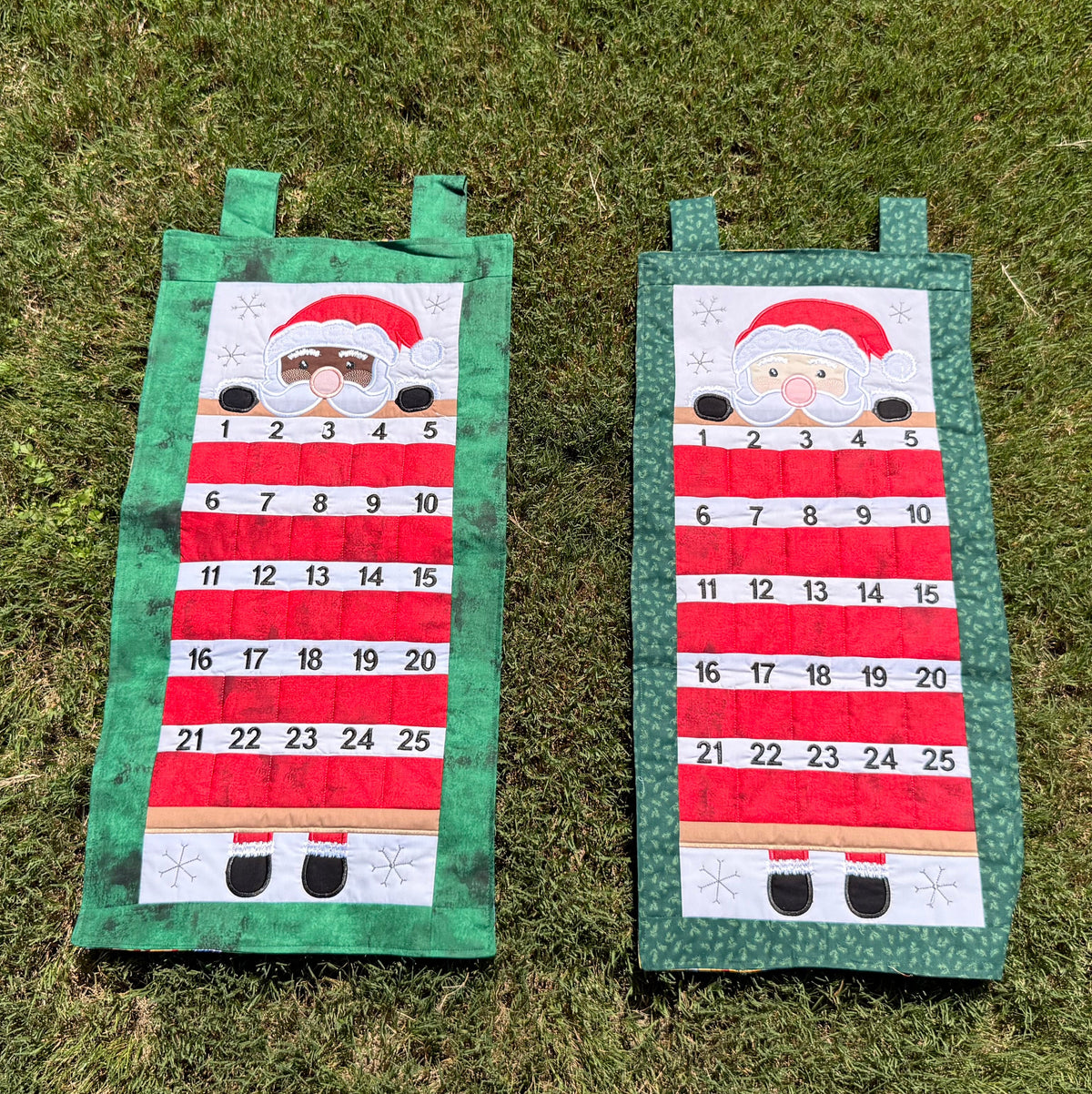 Two handmade fabric advent calendars on a green grass lawn. The calendar has Santa (one with light toned skin and the other with dark toned skin) peeking over the top section with numbered pockets in the lower section for stashing little goodies during the advent season.