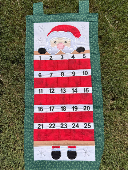 A handmade fabric advent calendar on a green grass lawn. The calendar has Santa (with light toned skin) peeking over the top section with numbered pockets in the lower section for stashing little goodies during the advent season.