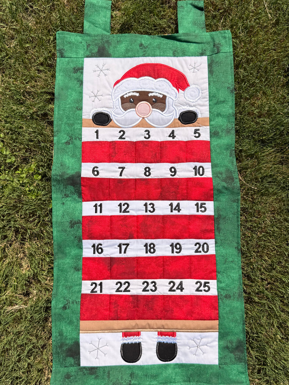 A handmade fabric advent calendar on a green grass lawn. The calendar has Santa (with dark toned skin) peeking over the top section with numbered pockets in the lower section for stashing little goodies during the advent season.