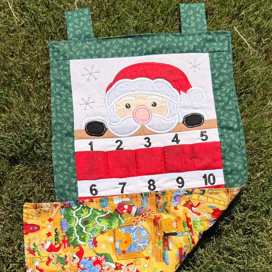 A handmade fabric advent calendar on a green grass lawn. The calendar has Santa (with light toned skin) peeking over the top section with numbered pockets in the lower section for stashing little goodies during the advent season. The bottom is flipped up so you can see the colorful fabric depicting Santa's workshop on the backside.