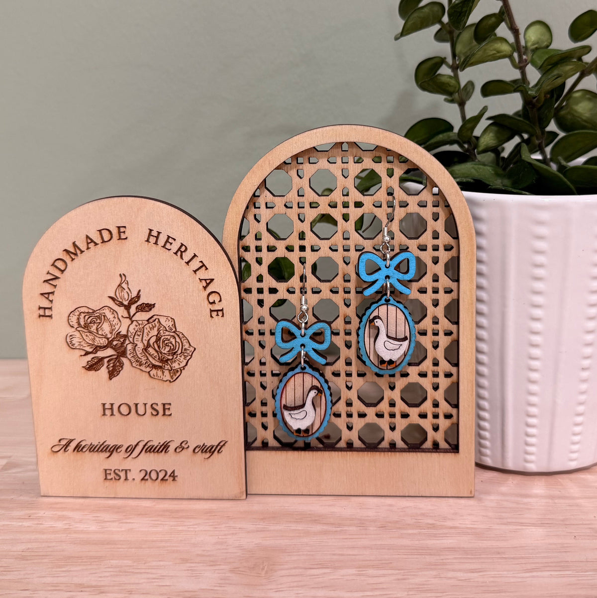 Handmade wood earrings displayed on a wooden stand. Each earring features a goose frame with a bow, crafted from birch wood with a gloss finish. Available in various colors including autumn red, mint green, and baby blue. The earrings come in different styles such as fish hooks, studs, and clip-ons, perfect for adding a whimsical touch to any outfit.