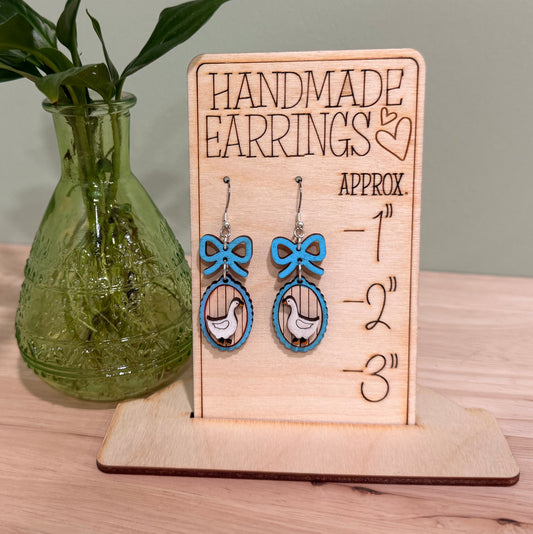 Handmade wood earrings displayed on a wooden stand. Each earring features a goose frame with a bow, crafted from birch wood with a gloss finish. Available in various colors including autumn red, mint green, and baby blue. The earrings come in different styles such as fish hooks, studs, and clip-ons, perfect for adding a whimsical touch to any outfit.