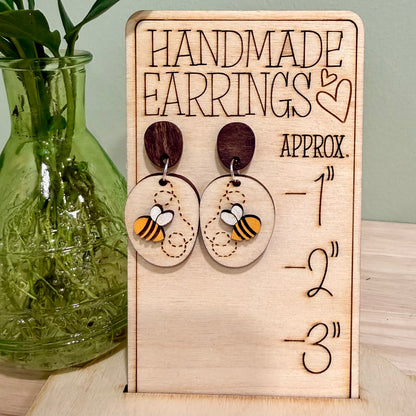 A pair of handmade wood earrings featuring a Busy Bee design. The earrings are crafted from birch wood with a bright yellow bee on a natural wood base with the bee's winding path engraved on it. They have hypoallergenic findings, available in stainless steel studs or clip-ons, or gold-plated clip-ons. The earrings are approximately 1 inch wide by 2 inches tall, with a satin finish and a total thickness of 0.5 inches. Perfect for nature-themed events and eco-friendly fashion.