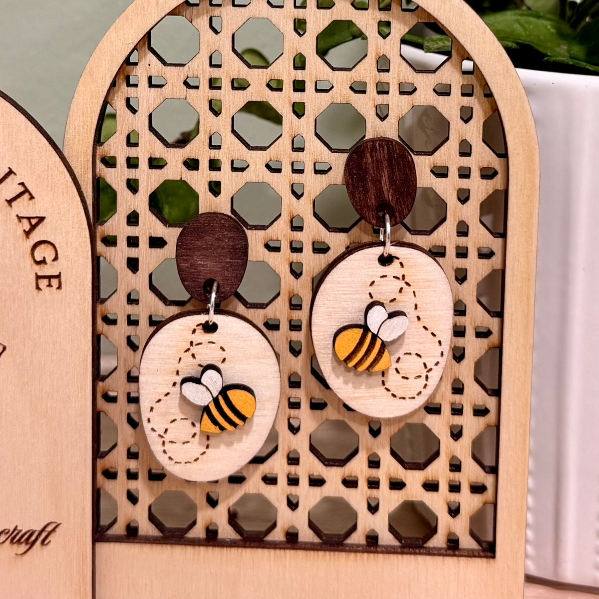 A pair of handmade wood earrings featuring a Busy Bee design. The earrings are crafted from birch wood with a bright yellow bee on a natural wood base with the bee's winding path engraved on it. They have hypoallergenic findings, available in stainless steel studs or clip-ons, or gold-plated clip-ons. The earrings are approximately 1 inch wide by 2 inches tall, with a satin finish and a total thickness of 0.5 inches. Perfect for nature-themed events and eco-friendly fashion.