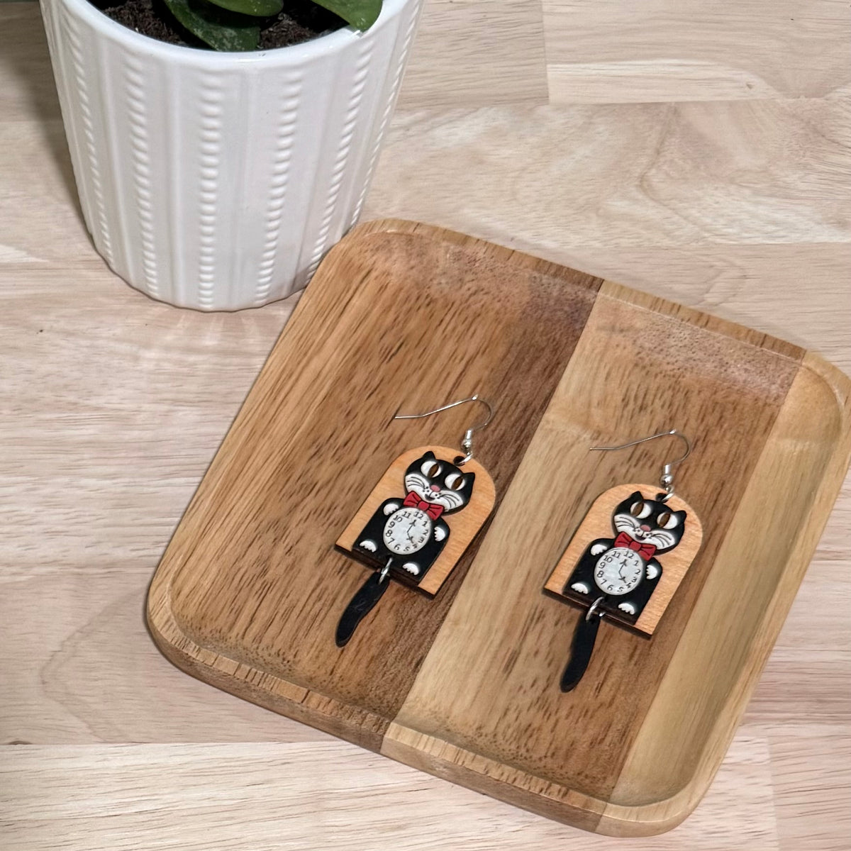 A pair of retro cat clock earrings handcrafted from birch wood, featuring a glossy finish and a whimsical black cat motif with a clock face. These earrings are available in fish hooks measuring 3.75 inches and clip-ons at 3.25 inches, crafted with hypoallergenic, nickel-free findings. The earrings offer a choice between gold-plated or stainless steel and allow for custom color requests. Displayed on a wooden stand, these earrings make a charming addition to any jewelry collection.