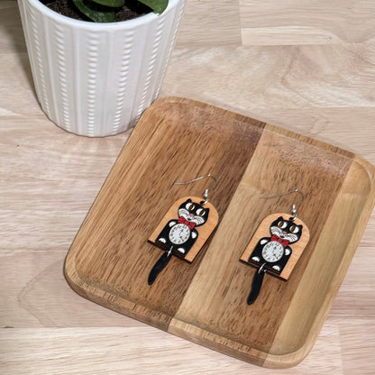 A pair of retro cat clock earrings handcrafted from birch wood, featuring a glossy finish and a whimsical black cat motif with a clock face. These earrings are available in fish hooks measuring 3.75 inches and clip-ons at 3.25 inches, crafted with hypoallergenic, nickel-free findings. The earrings offer a choice between gold-plated or stainless steel and allow for custom color requests. Displayed on a wooden stand, these earrings make a charming addition to any jewelry collection.
