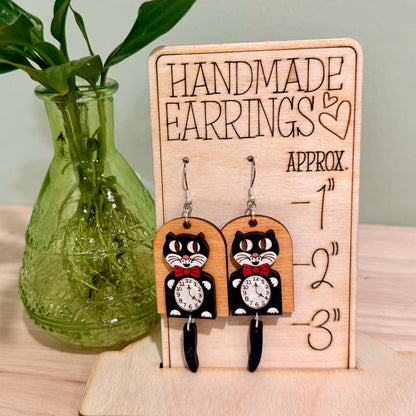 A pair of retro cat clock earrings handcrafted from birch wood, featuring a glossy finish and a whimsical black cat motif with a clock face. These earrings are available in fish hooks measuring 3.75 inches and clip-ons at 3.25 inches, crafted with hypoallergenic, nickel-free findings. The earrings offer a choice between gold-plated or stainless steel and allow for custom color requests. Displayed on a wooden stand, these earrings make a charming addition to any jewelry collection.
