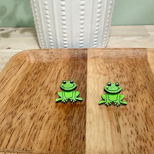 A pair of handmade wooden earrings featuring a smiling frog design, crafted from birch wood with a gloss finish. Available in bright and vintage green colors, these earrings offer hypoallergenic stainless steel and gold-plated options. The earrings are approximately 1 inch wide and 0.75 inches tall, suitable for nature lovers and those seeking unique, personalized jewelry pieces. Custom color options are available upon request, making these earrings a versatile accessory for various occasions.