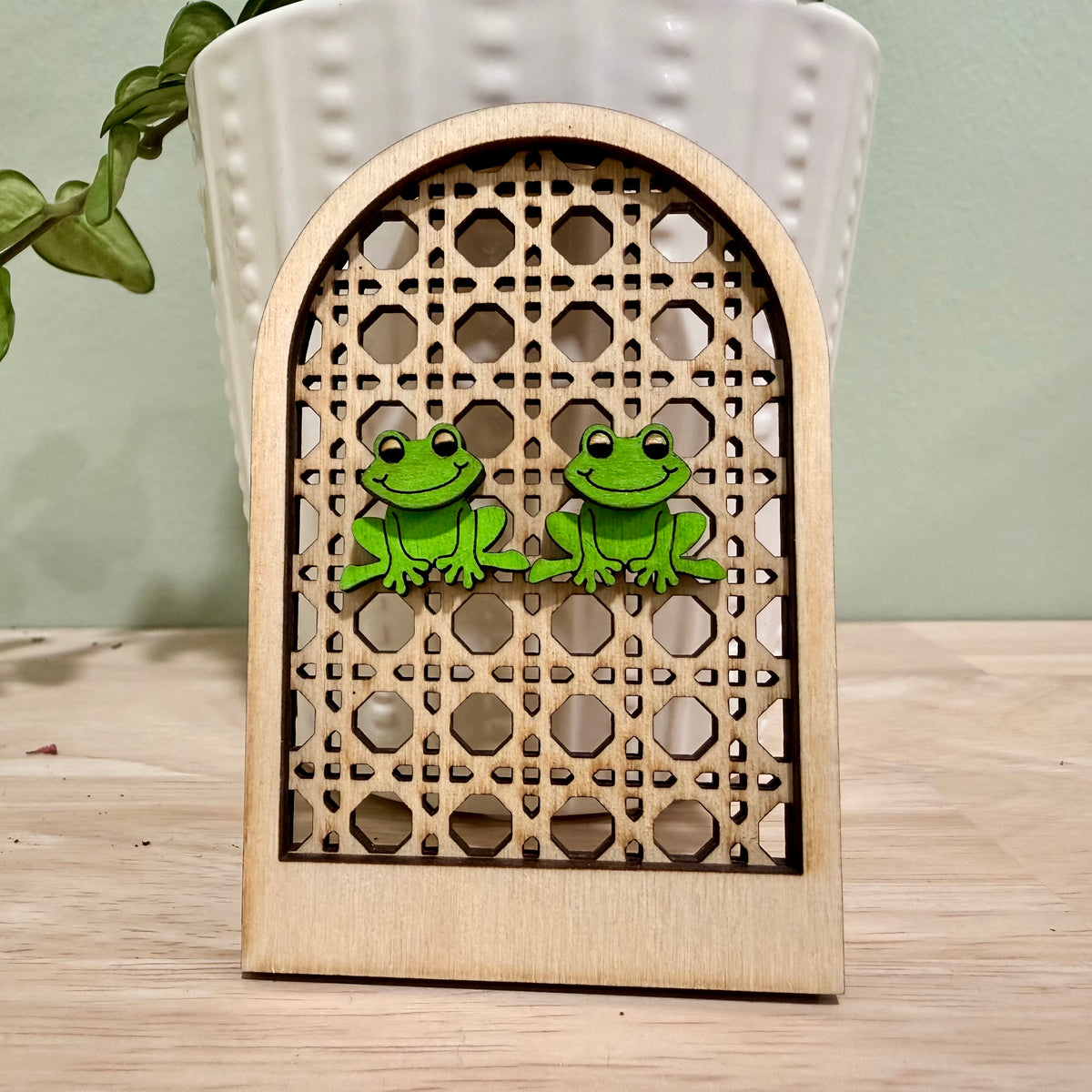 A pair of handmade wooden earrings featuring a smiling frog design, crafted from birch wood with a gloss finish. Available in bright and vintage green colors, these earrings offer hypoallergenic stainless steel and gold-plated options. The earrings are approximately 1 inch wide and 0.75 inches tall, suitable for nature lovers and those seeking unique, personalized jewelry pieces. Custom color options are available upon request, making these earrings a versatile accessory for various occasions.