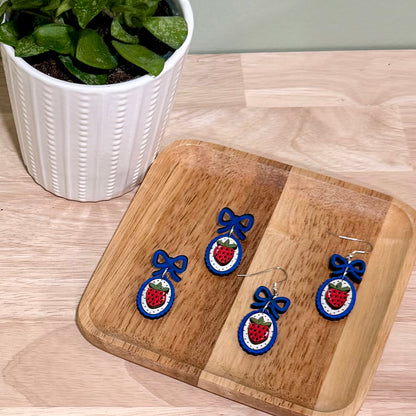 The image shows a pair of handmade earrings featuring a strawberry design with a coquette bow, crafted from birch wood. The earrings are displayed on a decorative wooden tray. These earrings highlight a gloss finish and are available in bright blue and dark blue, offering hypoallergenic findings with gold-plated or stainless steel options. The background features a green plant, adding a natural touch to the display.