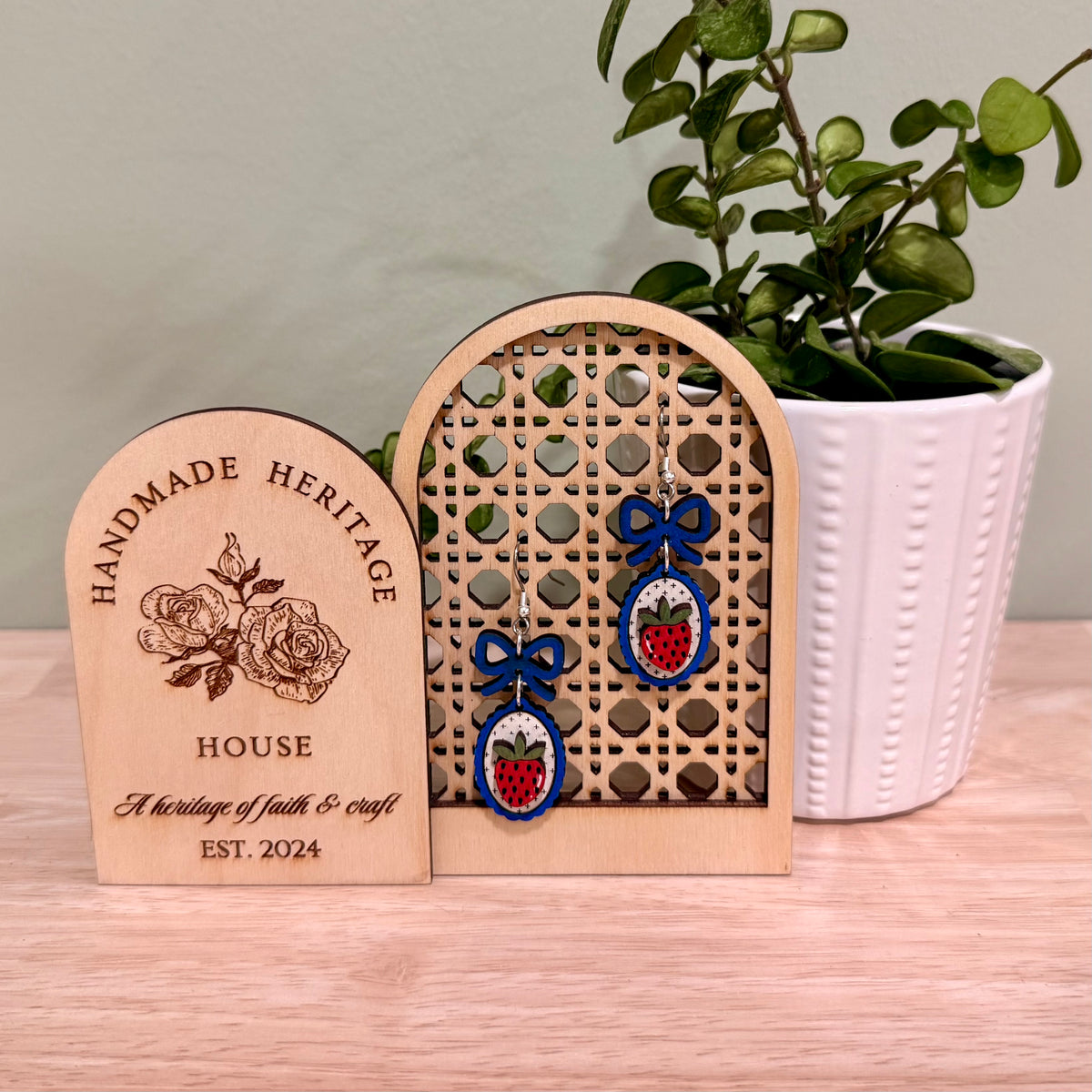 The image shows a pair of handmade earrings featuring a strawberry design with a coquette bow, crafted from birch wood. The earrings are displayed on a decorative wooden stand with floral engravings, labeled "HANDMADE HERITAGE HOUSE." These earrings highlight a gloss finish and are available in bright blue and dark blue, offering hypoallergenic findings with gold-plated or stainless steel options. The background features a green plant, adding a natural touch to the display.
