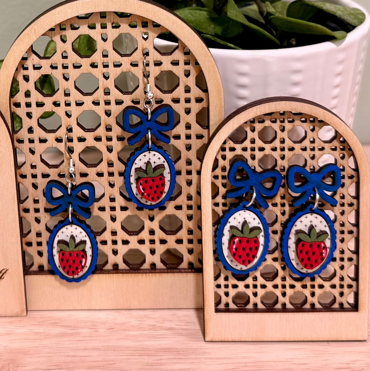 The image shows a pair of handmade earrings featuring a strawberry design with a coquette bow, crafted from birch wood. The earrings are displayed on a decorative wooden stand with floral engravings, labeled "HANDMADE HERITAGE HOUSE." These earrings highlight a gloss finish and are available in bright blue and dark blue, offering hypoallergenic findings with gold-plated or stainless steel options. The background features a green plant, adding a natural touch to the display.