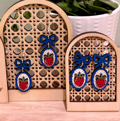 The image shows a pair of handmade earrings featuring a strawberry design with a coquette bow, crafted from birch wood. The earrings are displayed on a decorative wooden stand with floral engravings, labeled "HANDMADE HERITAGE HOUSE." These earrings highlight a gloss finish and are available in bright blue and dark blue, offering hypoallergenic findings with gold-plated or stainless steel options. The background features a green plant, adding a natural touch to the display.