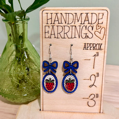 The image shows a pair of handmade earrings featuring a strawberry design with a coquette bow, crafted from birch wood. The earrings are displayed on a decorative wooden stand with heart engravings, labeled "HANDMADE EARRINGS." These earrings highlight a gloss finish and are available in bright blue and dark blue, offering hypoallergenic findings with gold-plated or stainless steel options. The background features a green plant, adding a natural touch to the display.