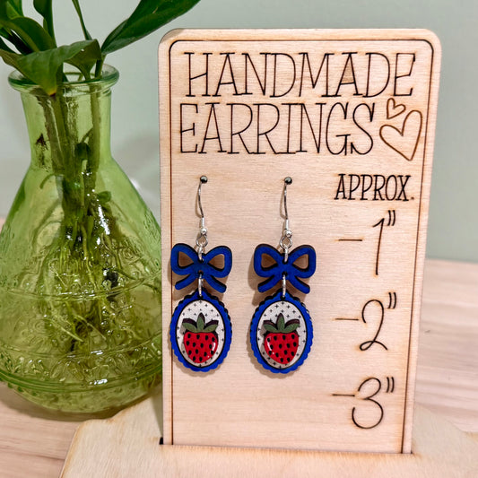 The image shows a pair of handmade earrings featuring a strawberry design with a coquette bow, crafted from birch wood. The earrings are displayed on a decorative wooden stand with heart engravings, labeled "HANDMADE EARRINGS." These earrings highlight a gloss finish and are available in bright blue and dark blue, offering hypoallergenic findings with gold-plated or stainless steel options. The background features a green plant, adding a natural touch to the display.