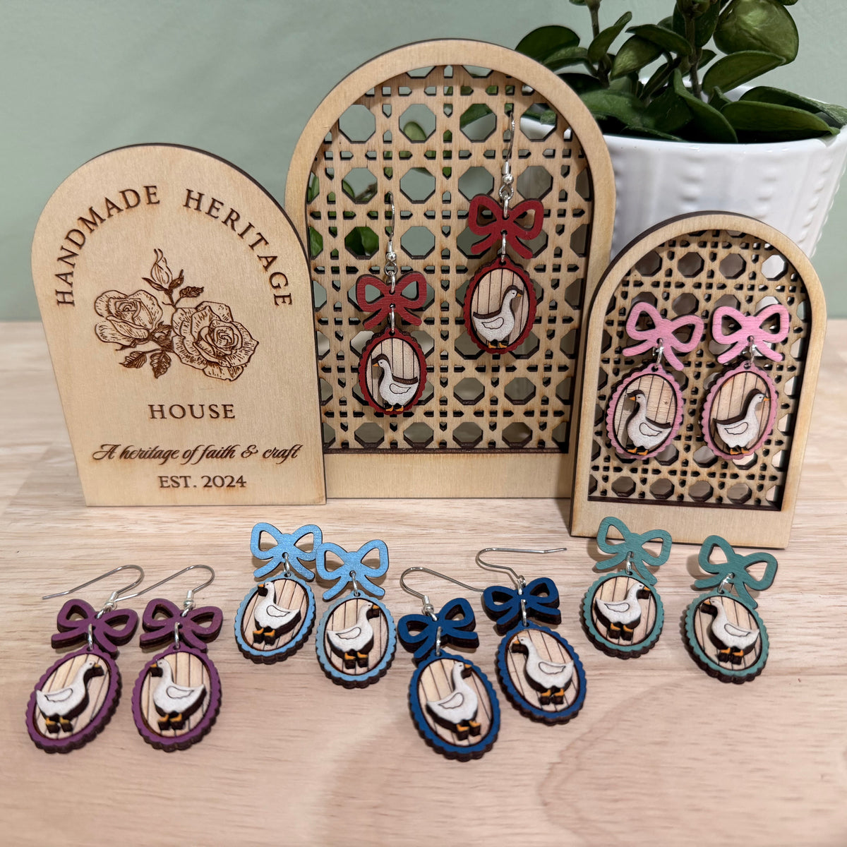 A collection of handmade wood earrings displayed on a wooden stand. Each earring features a goose frame with a bow, crafted from birch wood with a gloss finish. Available in various colors including autumn red, mint green, and baby blue. The earrings come in different styles such as fish hooks, studs, and clip-ons, perfect for adding a whimsical touch to any outfit.