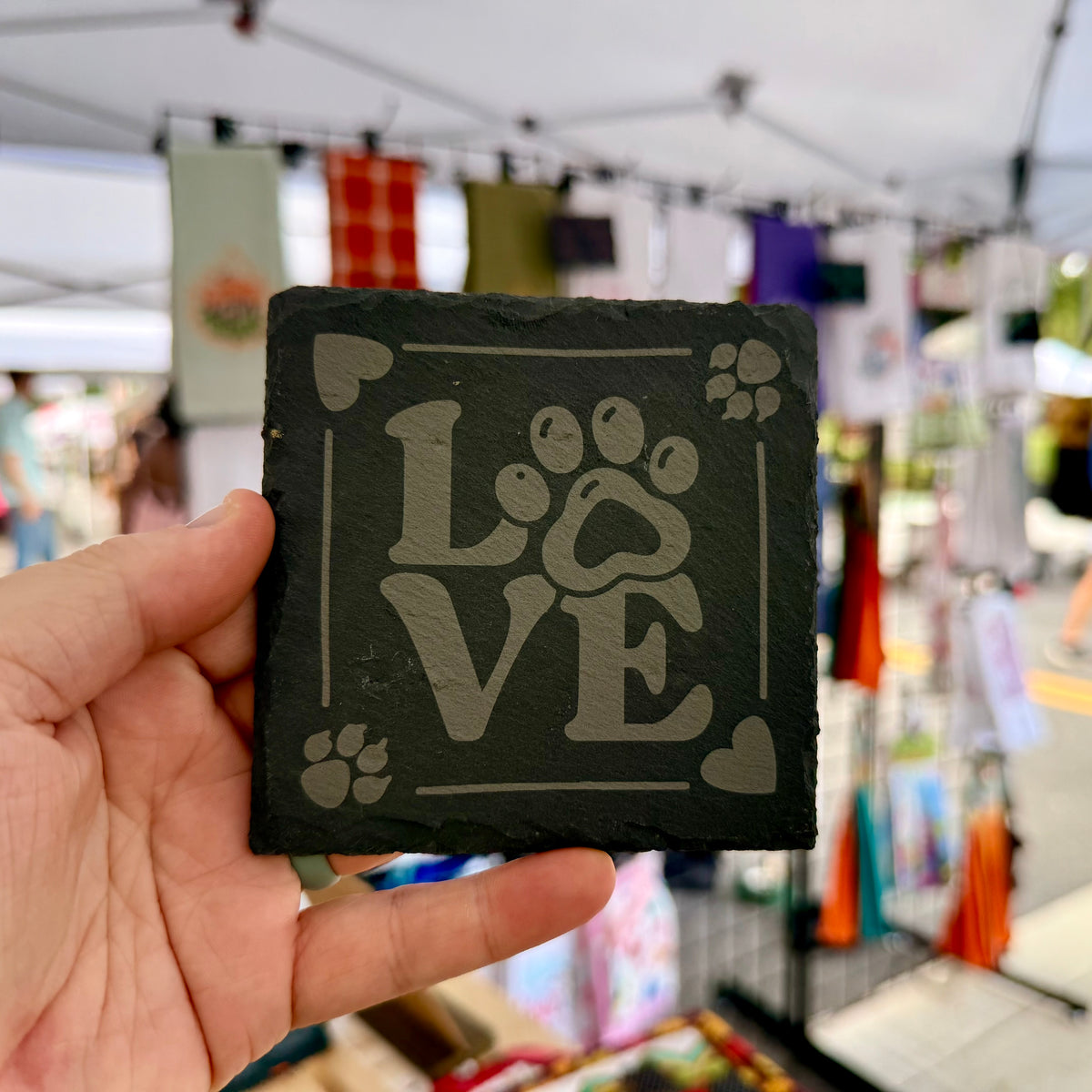 A close-up of a square slate coaster measuring 4 inches by 4 inches, featuring the word "LOVE" with the "O" represented by a paw print. The coaster is adorned with a border of paw prints and hearts, and is displayed against a craft fair background. This laser engraved coaster is ideal for pet lovers and adds a decorative touch to any home setting.