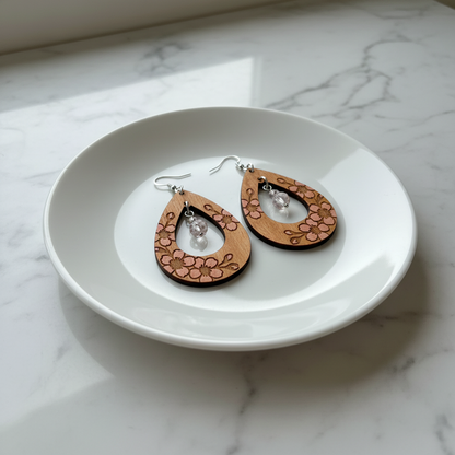 Pair of lightly stained wooden earrings with soft pink cherry blossom designs on a white plate with a marble background