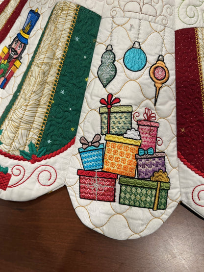 Detail view of a handcrafted white Christmas tree skirt featuring detailed embroidered Nutcracker Ballet characters around the border with detailed quilting in the center. Close up of Gifts.